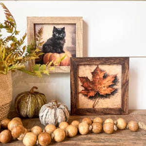 May include: Two framed pictures with fall decor. The first picture shows a black cat sitting on a pumpkin. The second picture shows a single brown maple leaf. The pictures are sitting on a wooden surface with a garland of brown wooden beads.