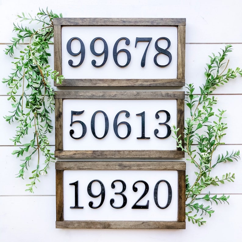 May include: Three rectangular wooden signs with white backgrounds and black numbers. The numbers are 99678, 50613, and 19320. The signs have dark brown wooden frames. Greenery is placed around the signs.