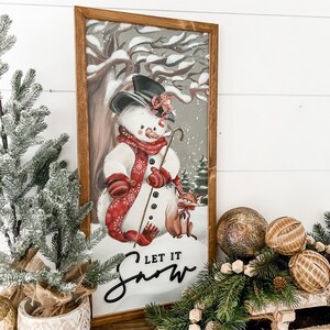 May include: A framed illustration of a snowman with a red scarf and mittens, holding a stick. The snowman is standing in a snowy forest with a fox beside it. The text "Let it Snow" is written below the snowman.