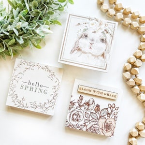 May include: Three wooden signs with spring-themed designs. The first sign features a wreath of leaves with the text "hello spring". The second sign features a watercolour illustration of a bunny wearing a flower crown. The third sign features a floral design with the text "bloom with grace".