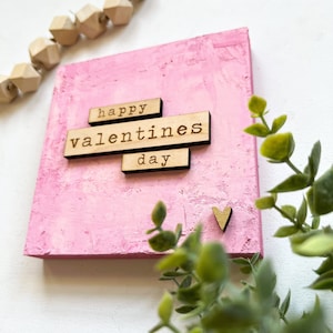May include: Pink canvas with the words "happy valentines day" in wooden letters. A small wooden heart is in the bottom right corner.