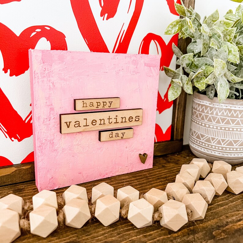 May include: A pink wooden sign with the words "happy valentines day" in wooden letters. The sign is sitting on a wooden surface with a wooden bead garland in front of it.