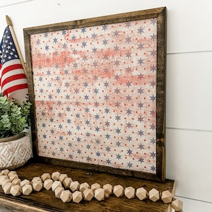 May include: A framed wood sign with a distressed American flag design featuring red, white, and blue stars on a white background. The sign is hanging on a white wall.