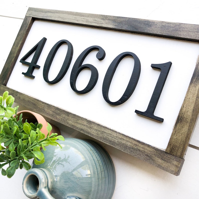 May include: A rectangular wooden sign with the numbers "40601" in black, mounted on a white background. The sign has a dark gray frame. Decorative items, including a small plant in a terracotta pot and a blue ceramic jug, are in the foreground.