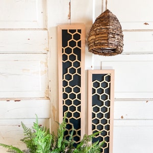 May include: Two wooden framed wall decor pieces with a black background and gold honeycomb pattern. The frames are a light wood color. A woven wicker pendant light hangs above the frames.