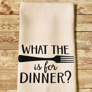 Puede incluir: Toalla de cocina blanca con texto negro que dice "What the is for dinner?" con un gráfico de tenedor negro.