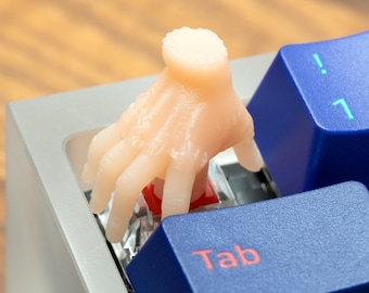May include: A close-up of a keyboard with a flesh-colored hand pressing down on the "Tab" key. The hand is made of a soft, pliable material and is designed to look like a human hand.
