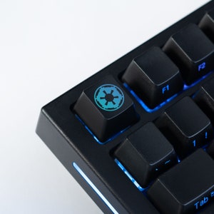 May include: Close-up of a black mechanical keyboard with blue backlighting. The keycap features a teal and blue Imperial symbol. Other keys are visible, including F1, F2, and Tab.