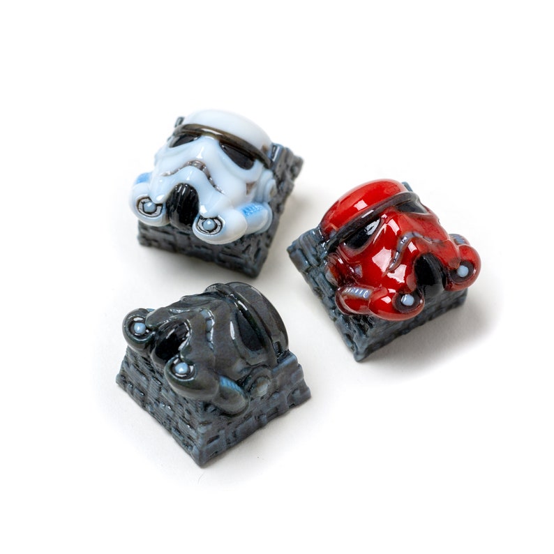Op de afbeelding: Drie Star Wars Stormtrooper-toetsen, elk met een helm in een andere kleur: wit, rood en zwart. De toetsen zijn gemaakt van hars en hebben een gedetailleerd beeldhouwwerk van de Stormtrooper-helm. De toetsen zijn gemonteerd op een grijze, bakstenen basis.