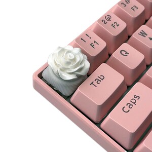Pu&ograve; includere: Un copritasto a forma di rosa bianca per una tastiera meccanica. Il copritasto &egrave; realizzato in plastica bianca liscia e presenta un delicato design a forma di rosa. Il copritasto &egrave; posto su una tastiera rosa.