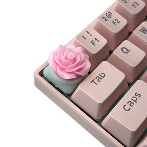 Pu&ograve; includere: Un copritasto rosa a forma di rosa per una tastiera meccanica. Il copritasto &egrave; realizzato in resina e presenta un design di rosa dettagliato. Il copritasto &egrave; su una tastiera rosa con i tasti "Tab" e "Caps" visibili.