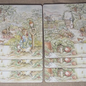 May include: A set of rectangular placemats featuring a detailed watercolor illustration of Peter Rabbit in a vegetable garden. The scene includes a rabbit wearing a blue jacket, various garden vegetables, a small bird, and a shovel handle.