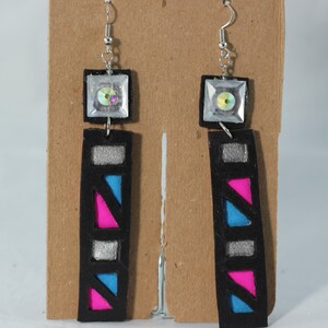 May include: Geometric dangle earrings with a black base, featuring silver, pink, and blue accents. Each earring has a square top with a clear, iridescent gem. The design is modern and eye-catching.