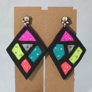 May include: A pair of diamond-shaped earrings with a black outline and vibrant, geometric segments. The segments are filled with neon pink, green, teal, and silver, and are embellished with small, sparkling rhinestones. The earrings have a silver post with iridescent crystals.