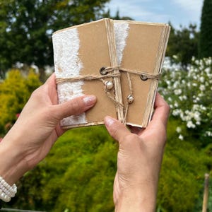 May include: Two small, handmade journals with kraft paper covers and white textured accents. The journals are tied together with twine and adorned with pearl beads. The background is a blurred view of green foliage and a blue sky.