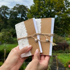 May include: Two small, handmade notebooks with white textured paper and brown covers are tied together with twine and small silver-colored charms. The notebooks are held in front of a blurred green background.