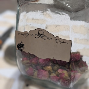 May include: A glass jar filled with dried rose buds. The jar has a label with a skull and crossbones and the text "Est. 1876".