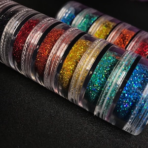 Op de afbeelding: Een set van acht kleine potjes gevuld met kleurrijke glitter. De potjes zijn gestapeld in een regenboogpatroon, met zwarte glitter onderaan en blauwe glitter bovenaan.