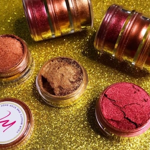 Puede incluir: Cuatro pequeños frascos de polvo de pigmento metálico en tonos cobre, oro y rojo. Los frascos están sobre un fondo de purpurina dorada. La etiqueta de un frasco dice "Your Beauty Elixir End Stop Shop".