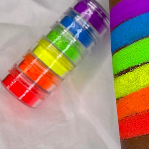 May include: A set of six small jars filled with neon colored glitter. The jars are stacked on top of each other and the glitter is visible through the clear jars. The glitter is in the colors red, orange, yellow, green, blue, and purple.