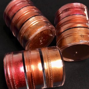 Puede incluir: Tres recipientes de plástico transparente con múltiples capas de polvo de pigmento metálico brillante. Los polvos son tonos de rojo, naranja y dorado.