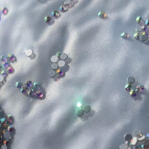 Può includere: Primo piano di strass iridescenti sparsi su un tessuto azzurro chiaro. Gli strass sono rotondi e sfaccettati, riflettendo uno spettro di colori. Il tessuto ha una consistenza morbida e leggermente stropicciata, creando un effetto delicato e scintillante.