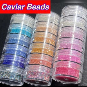 May include: Three clear plastic containers filled with colorful caviar beads. The beads are arranged in layers of blue, gold, and pink hues. The text "Caviar Beads" is displayed in a red box at the top of the image.