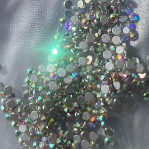 Op de afbeelding: Een close-up van een verzameling iriserende strass-steentjes verspreid over een lichtgrijze ondergrond. De strass-steentjes zijn rond met een platte achterkant en reflecteren een spectrum aan kleuren, waaronder groen, paars en goud. Het effect is sprankelend en decoratief.