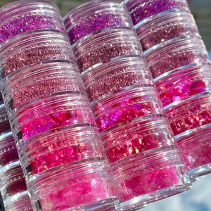 May include: A set of six clear plastic jars filled with various shades of pink glitter. The jars are stacked on top of each other, creating a tower of glitter.