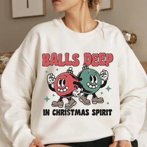 Pode incluir: Camisola branca com um gráfico de Natal. O gráfico apresenta dois ornamentos de desenho animado com rostos, um vermelho e outro verde, e o texto "Balls Deep in Christmas Spirit."