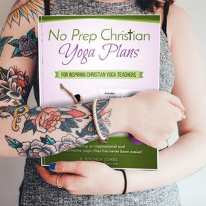 May include: A pink and green book titled "No Prep Christian Yoga Plans" for inspiring Christian yoga teachers. The book includes 30 Bible-based yoga plans. The book is held by a person with tattoos on their arms.