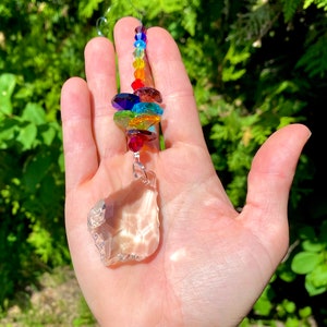 May include: A decorative suncatcher with a large, clear, irregular-shaped crystal at the bottom. It features a string of colourful, faceted crystals in a rainbow of hues, including red, orange, yellow, green, blue, and purple.