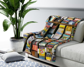 Pode incluir: Uma manta decorativa sobre um sofá branco, com uma estampa colorida de estantes cheias de livros. O design da manta inclui várias capas de livros com títulos e cores. Uma grande planta em vaso está à esquerda.