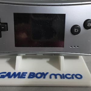 May include: Silver Game Boy Micro handheld game console on a white stand with the words "GAME BOY micro" in blue. The console has a black screen, a directional pad, and two buttons. The stand has two support legs.