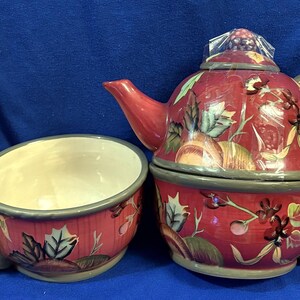 May include: A ceramic tea set consisting of a teapot and a matching mug, both featuring a red base color with a painted pattern of green leaves, brown acorns, and small berries. The teapot has a gray handle and a lid, while the mug has a gray handle.