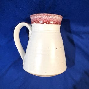 May include: White ceramic mug with a flared body and a curved handle. The rim of the mug is a deep red color, contrasting with the white body. The mug has a handmade appearance, suggesting it is a unique piece of pottery.