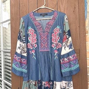 Op de afbeelding: Een blauwe spijker-tuniek met een V-hals en gedetailleerd roze en rood bloemenborduursel. Het kledingstuk heeft lange klokmouwen met veelkleurige patroonpanelen en een geplooide zoom met diverse bloemrijke en geometrische stoffen prints.