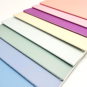 May include: A stack of seven rectangular notebooks with solid-colored covers in shades of peach, pink, purple, cream, light green, sage green, and light blue. Each notebook features a visible stitched spine along the left edge of the cover.