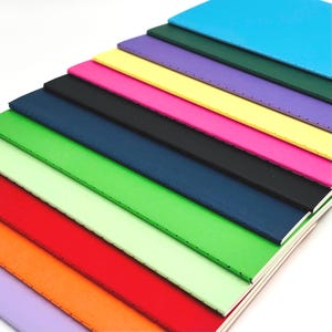 Può includere: Una pila di undici quaderni rettangolari disposti in fila diagonale. Le copertine presentano una varietà di colori a tinta unita tra cui viola chiaro, arancione, rosso, verde chiaro, verde brillante, blu scuro, nero, rosa, giallo, viola, verde scuro e blu.