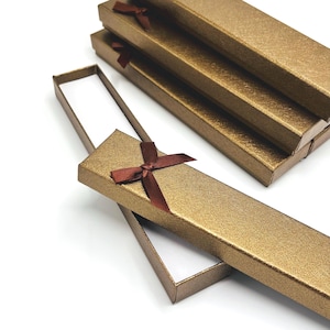 May include: A collection of rectangular jewelry gift boxes with a textured metallic gold finish. Each box features a dark brown satin ribbon tied in a bow on the lid. The boxes are arranged in a stack, with one box lid partially removed.
