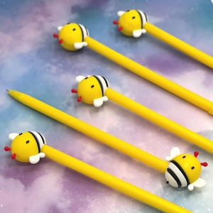 Pode incluir: Canetas amarelas com enfeites em forma de abelha. Os enfeites de abelha são amarelos com listras pretas, asas brancas e antenas vermelhas. As canetas estão dispostas sobre um fundo colorido e desfocado.