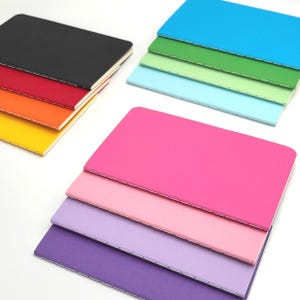 May include: A collection of twelve rectangular notebooks arranged in three stacks. The notebooks feature solid-colored covers in black, red, orange, yellow, bright blue, green, light green, light blue, hot pink, light pink, lavender, and purple.