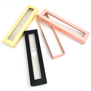 Puede incluir: Cuatro cajas rectangulares para bolígrafos en colores pastel: amarillo, negro y rosa. Cada caja tiene una ventana rectangular transparente en la tapa, que revela el interior. Las cajas están abiertas y cerradas, mostrando su diseño y uso potencial.