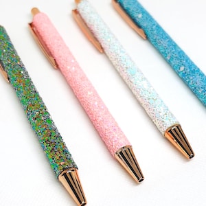 Könnte beinhalten: Vier Glitzerstifte in Grün, Rosa, Weiß und Blau. Jeder Stift hat einen goldfarbenen Clip und eine Spitze. Die Stifte sind mit Glitzer bedeckt, was einen funkelnden Effekt erzeugt. Die Stifte sind diagonal angeordnet.