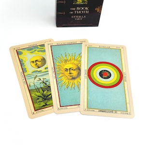Può includere: Tre carte dei tarocchi del mazzo The Book of Thoth Etteilla sono esposte. La carta a sinistra mostra una luna e piante etichettate Les Plantes. La carta centrale presenta un sole etichettato La Lumiere. A destra sono visibili cerchi concentrici.