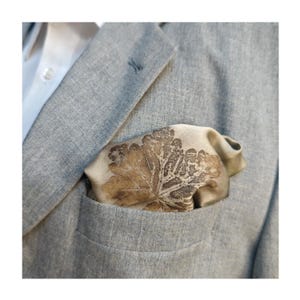 May include: A light gray blazer with a white shirt peeking out. The blazer's pocket holds a beige pocket square with a brown leaf design. The pocket square is folded to show the leaf pattern.