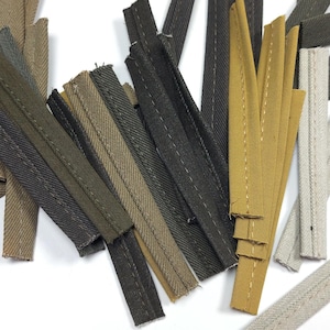 May include: Assortment of fabric zipper strips in various colors, including olive green, black, tan, and mustard yellow. The strips are arranged on a white surface, showcasing their different shades and textures. Ideal for sewing and crafting projects.