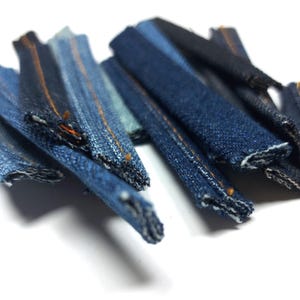 Könnte beinhalten: Eine Sammlung von Denim-Stoffstreifen mit rohen Kanten und orangefarbener Naht. Die Streifen sind fächerförmig angeordnet und in verschiedenen Blautönen gehalten.