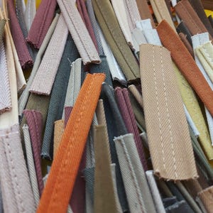 May include: A collection of fabric piping strips in various colors including orange, brown, olive green, beige, and grey. Each strip features a folded edge with visible stitching running along the length of the fabric, creating a textured appearance.