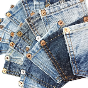 Może przedstawiać: Zbliżenie na stos skrawków tkaniny dżinsowej z widocznymi szwami i metalowymi nitami. Tkanina jest w różnych odcieniach niebieskiego i ma zużyty, postarzony wygląd.